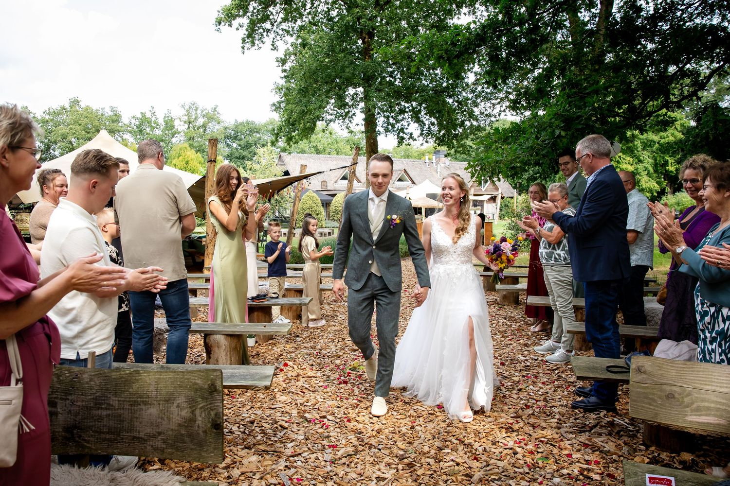 Gasten lopen langs een houten pad bij de uitgang van de huwelijksceremonie, terwijl het bruidspaar feestviert.