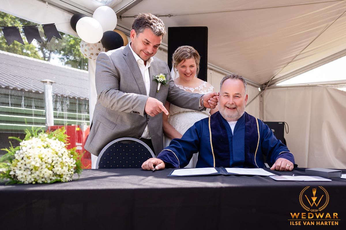 Couple signing wedding documents with officiant at outdoor ceremony under white tent with floral arrangements.