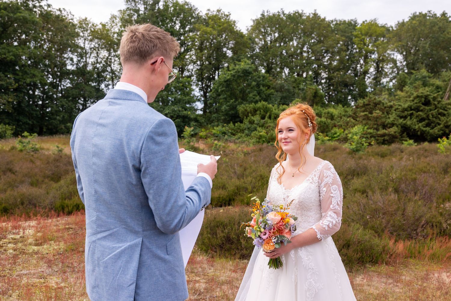 A couple exchanging vows in a rustic outdoor setting with heather and trees in the background.