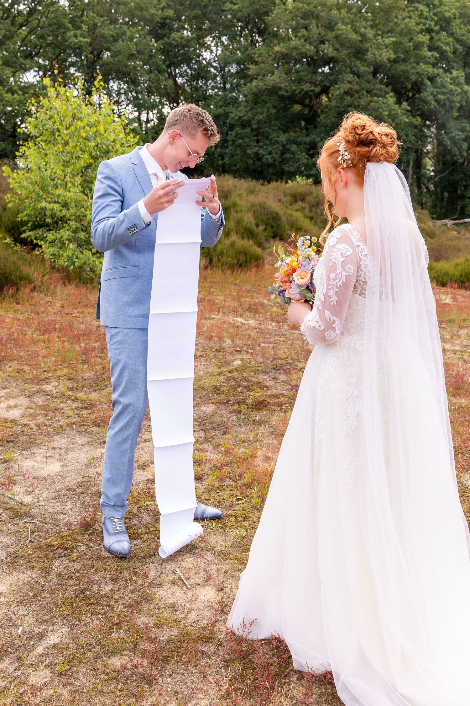 A wedding ceremony moment as the groom reads from a long scroll while the bride holds her bouquet.