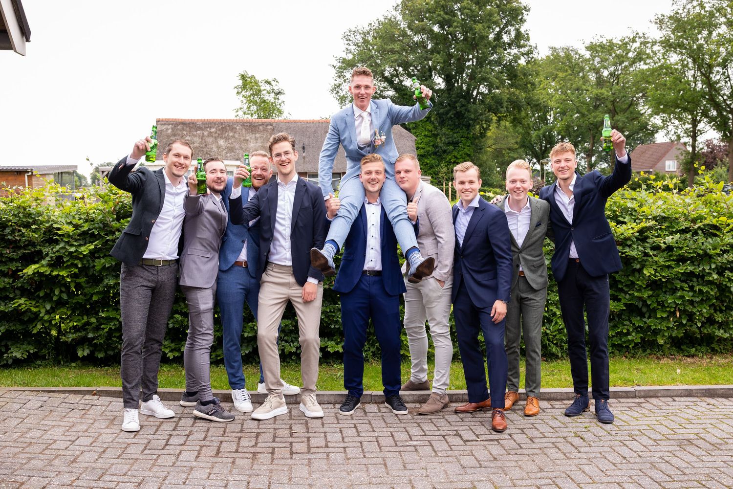 Groomsmen celebrate with energetic poses on brick pathway with garden backdrop.