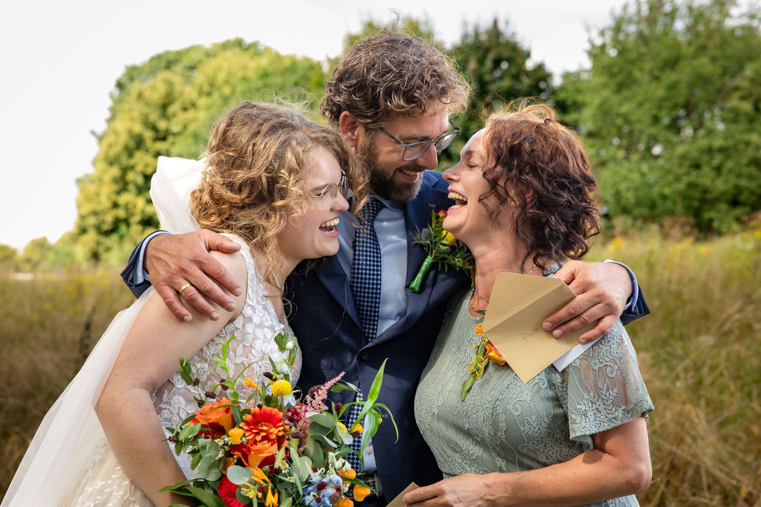 Wedding party embraces and laughs together in a sunny outdoor setting with natural greenery.