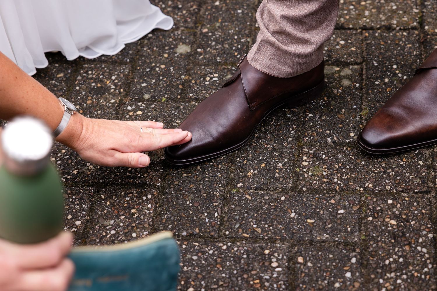 Close-up of formal shoes and a white wedding dress hem on textured ground.