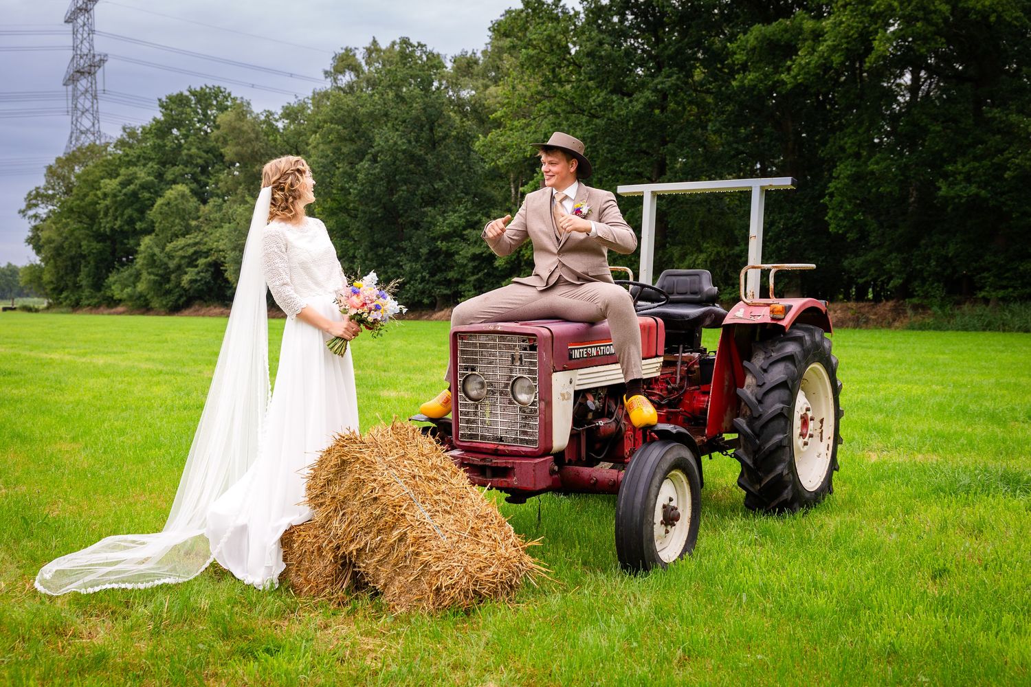 A rustic wedding photo featuring a vintage red tractor in a green field with hay bales.
