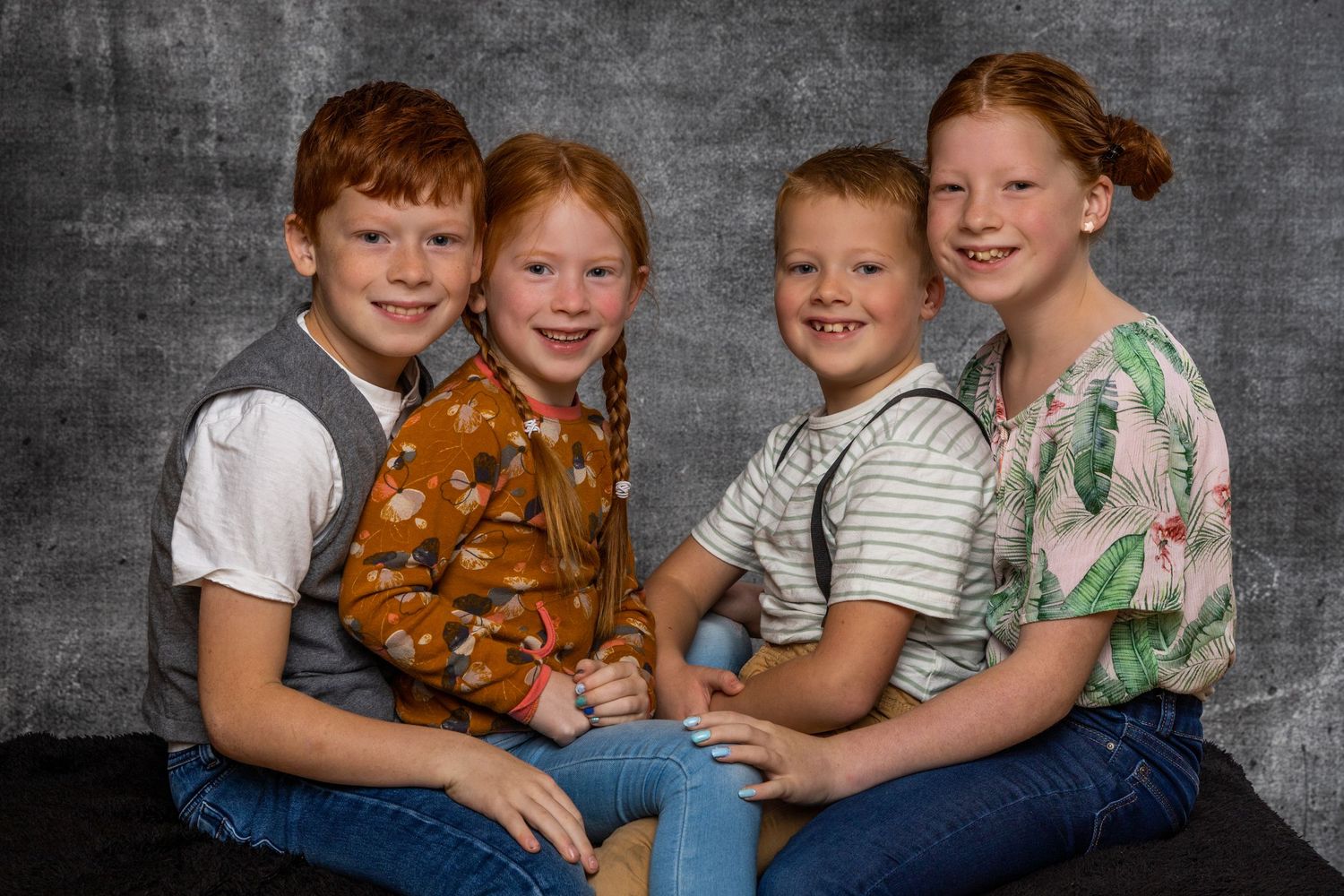 A group of siblings sit close together on a gray backdrop wearing casual clothing and smiling at the camera.