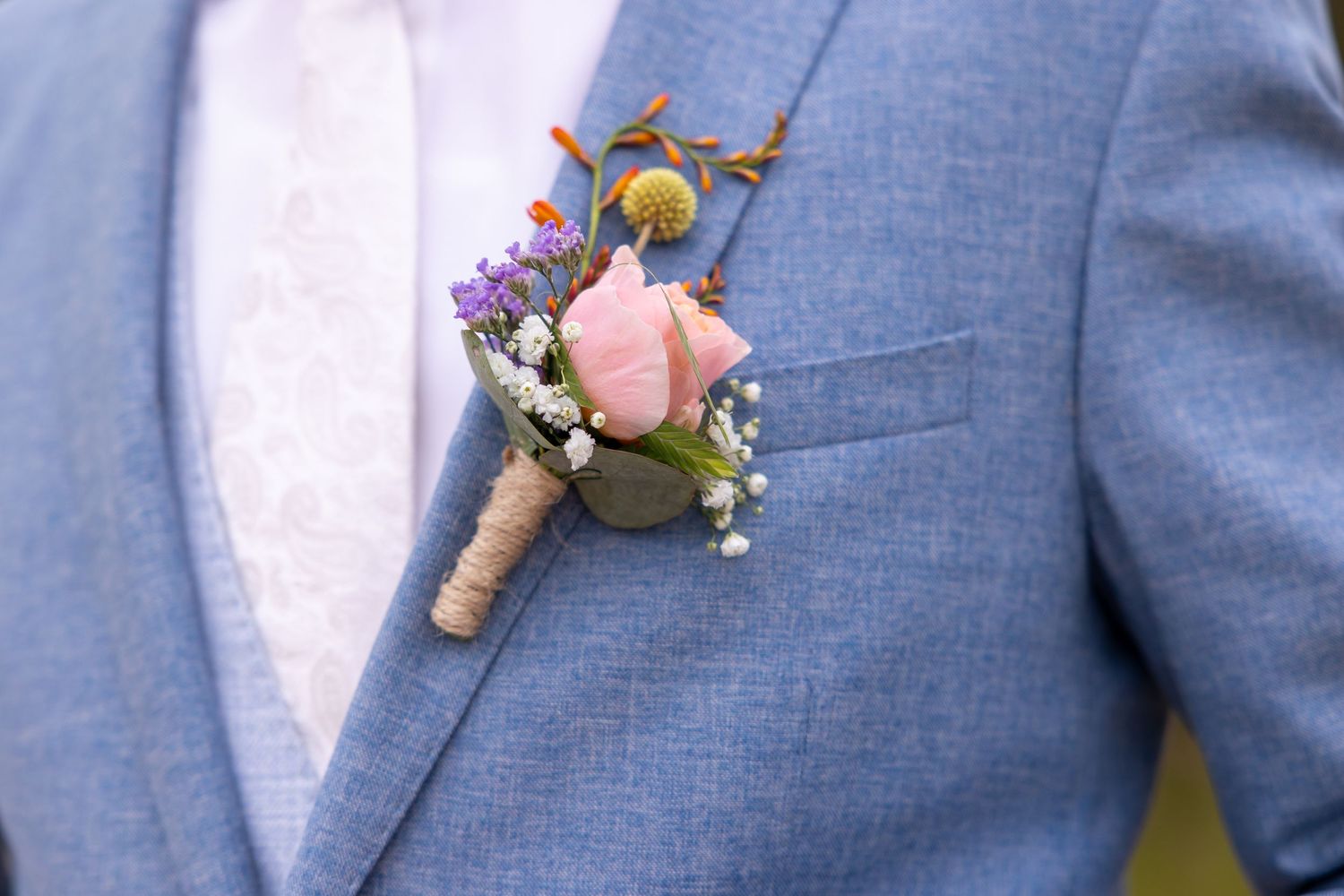 A delicate boutonniere with pink and purple flowers pinned to a light blue suit jacket.