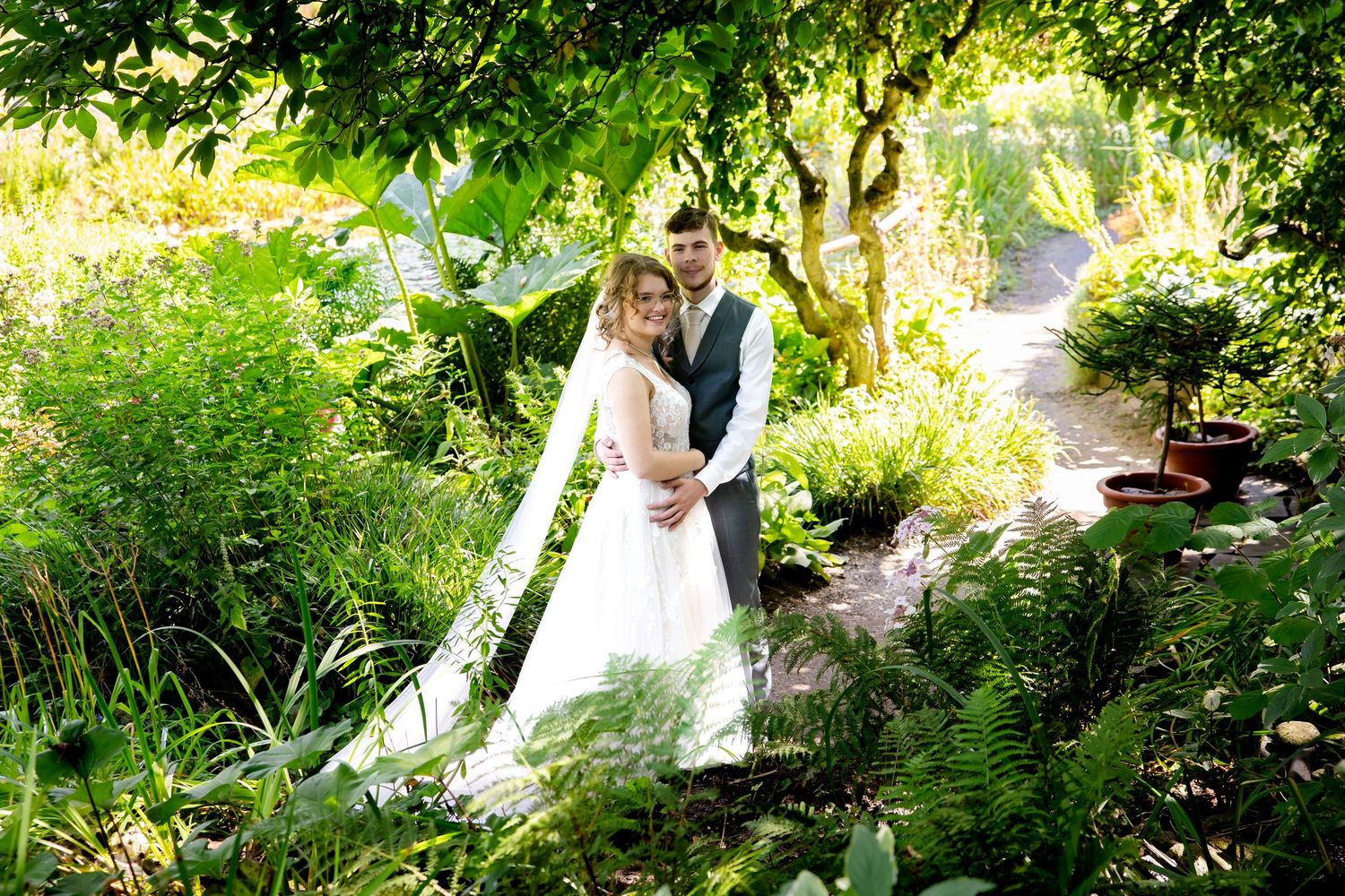 Wedding couple poses in lush garden setting with natural sunlight filtering through trees.