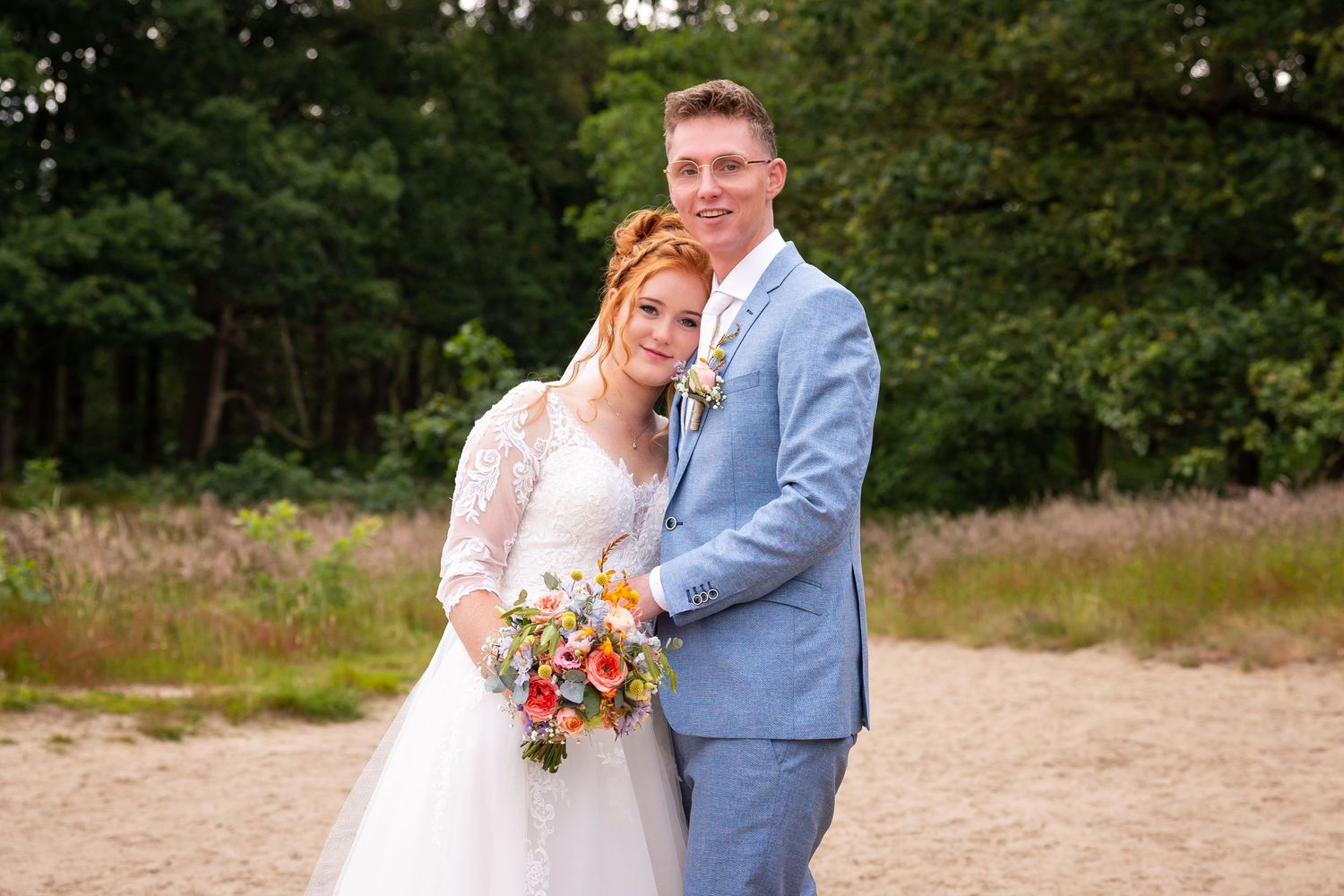 A romantic wedding portrait of a couple in a light blue suit and white dress against a natural backdrop.