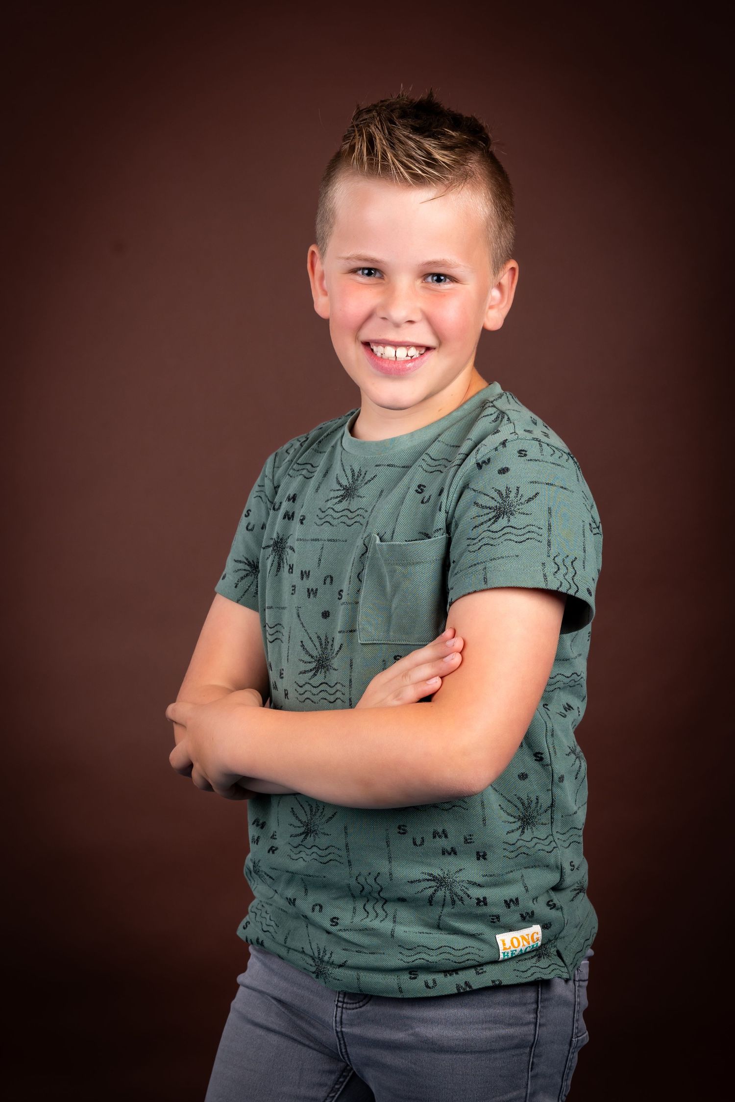 A young child poses confidently in a green patterned t-shirt and jeans against a dark brown studio background.