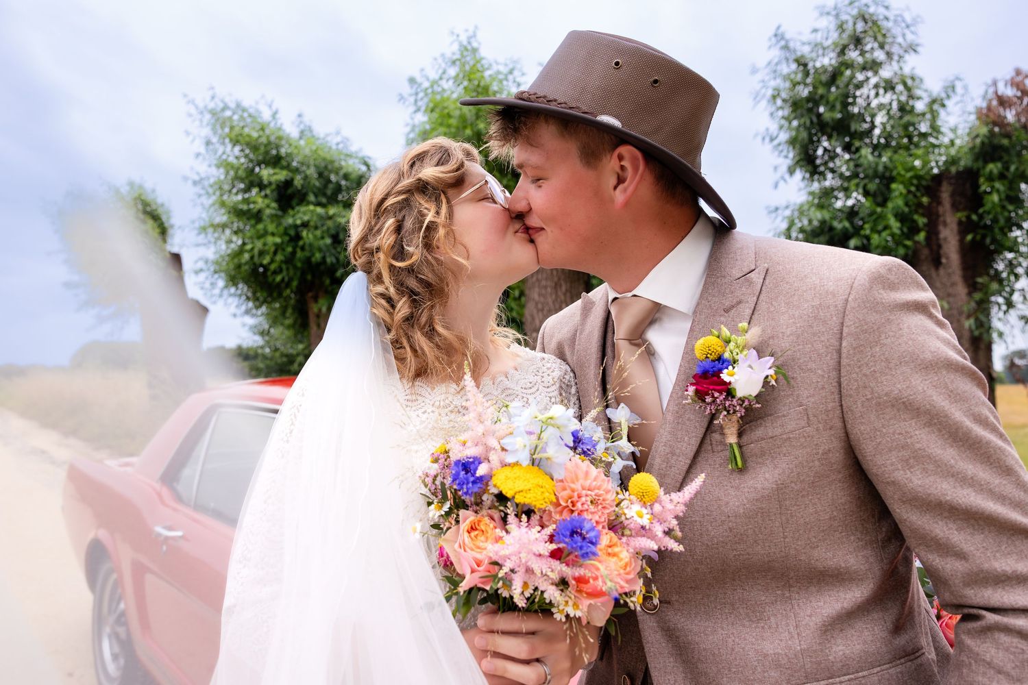 A romantic wedding moment captured with colorful wildflower bouquet and vintage car in background.