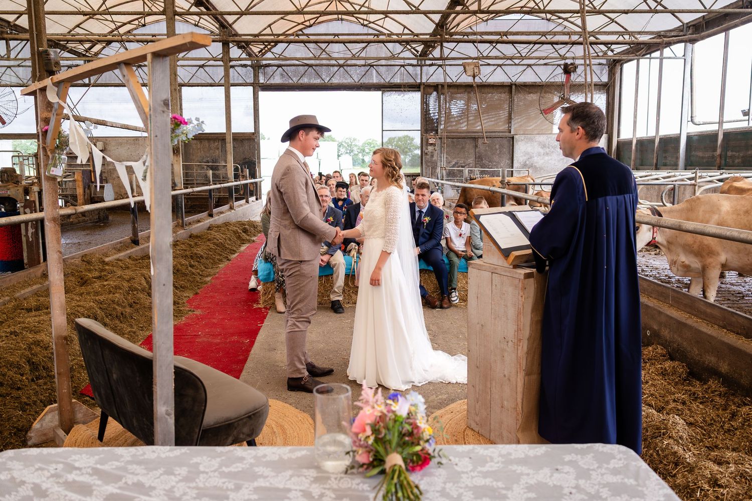 Wedding ceremony conducted in a barn with red carpet and farm animals visible in the background.