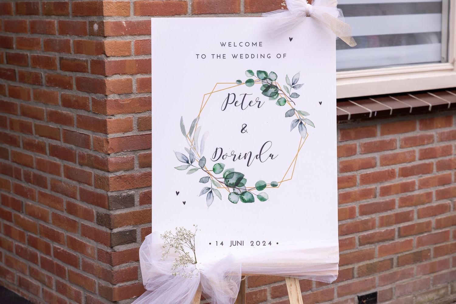 Elegant wedding welcome sign with floral design on white board against brick wall.