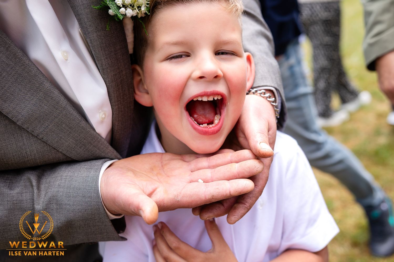 Joyful child laughing with mouth wide open at a wedding, wearing a floral crown, held by an adult in a gray suit.