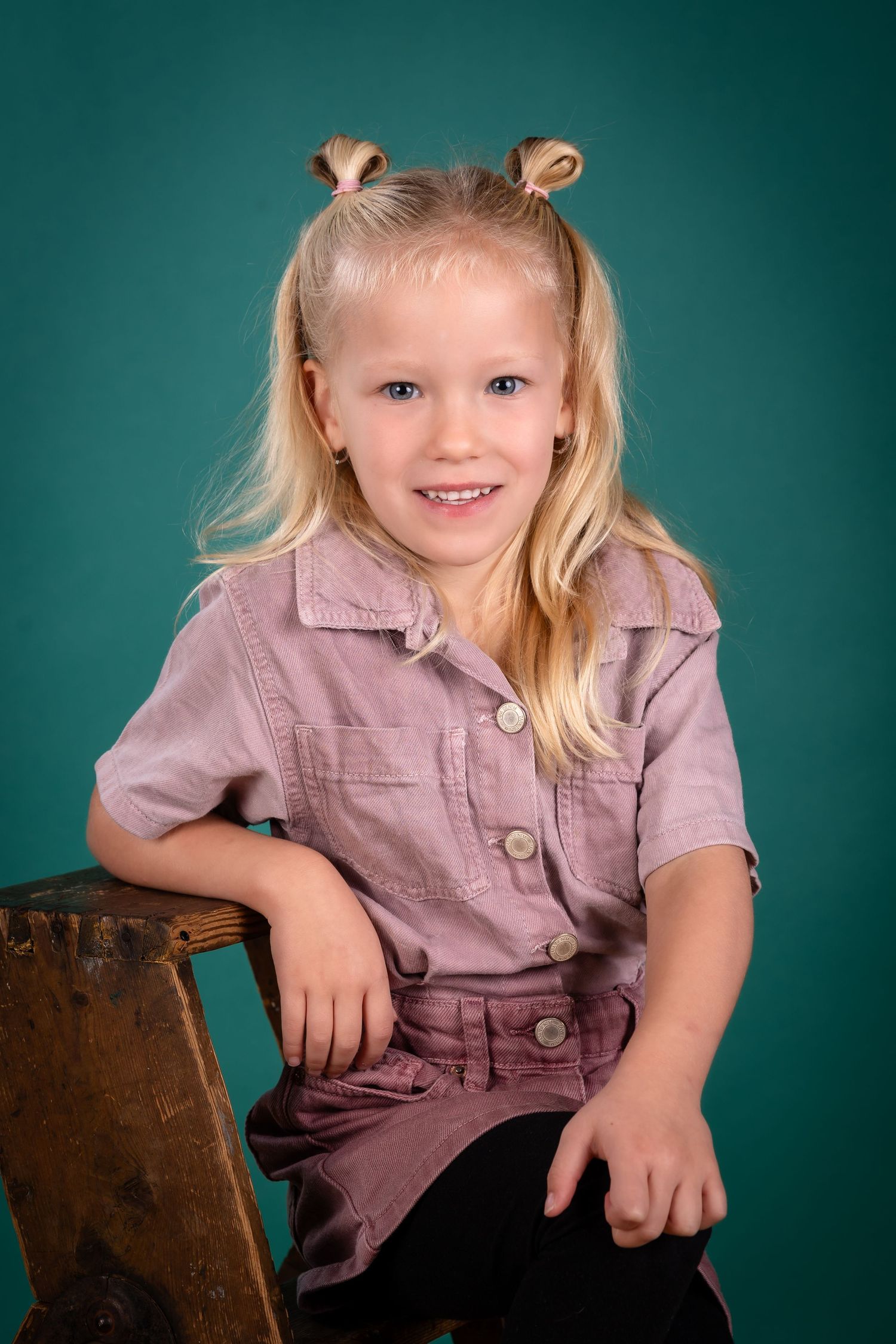 Young child in mauve button-up shirt poses on wooden furniture against teal backdrop in studio portrait setting.