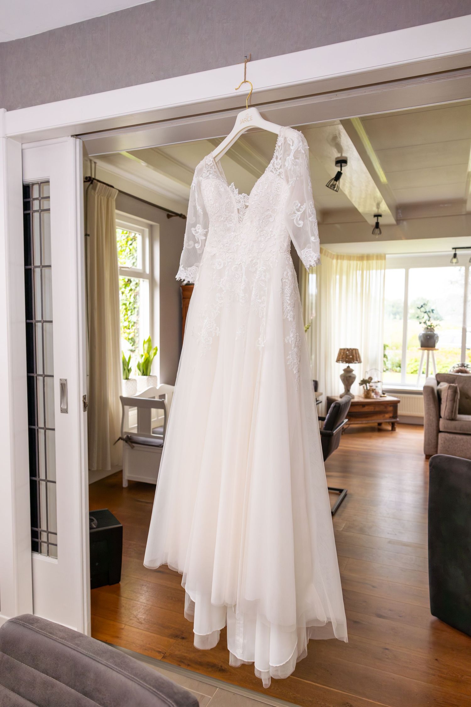 White lace wedding dress hanging in doorway of sunlit room.