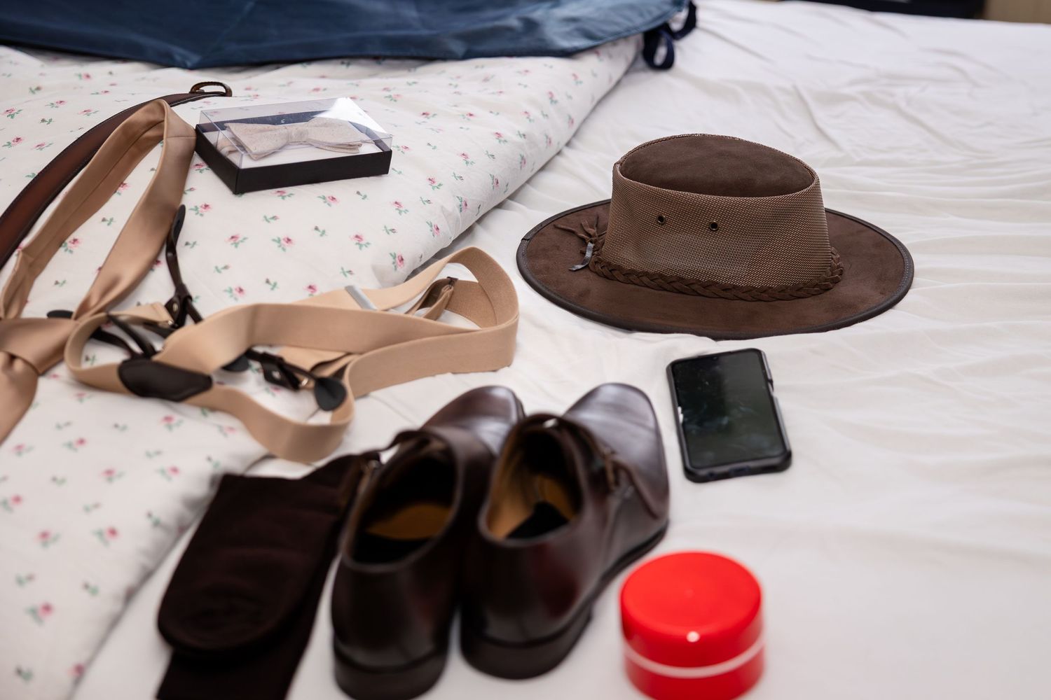 Wedding accessories including brown dress shoes, a hat, and formal attire laid out on a white surface.