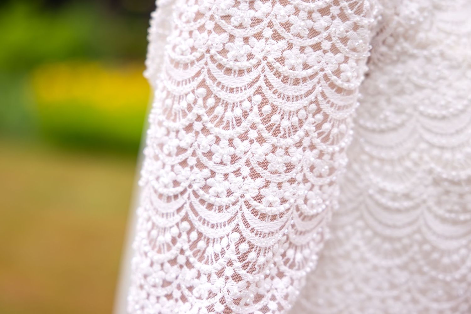 Close-up detail of delicate white lace pattern from a wedding dress against a green outdoor background.