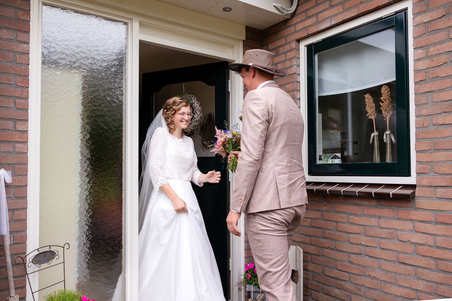 A couple stands in the doorway of a brick house on their wedding day.