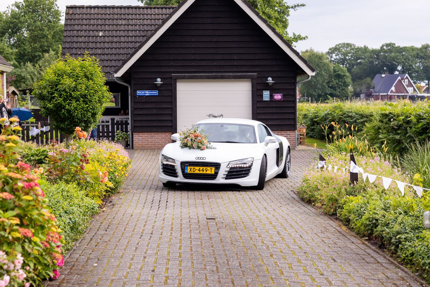 A white Audi R8 sports car parked in front of a black wooden garage surrounded by gardens.