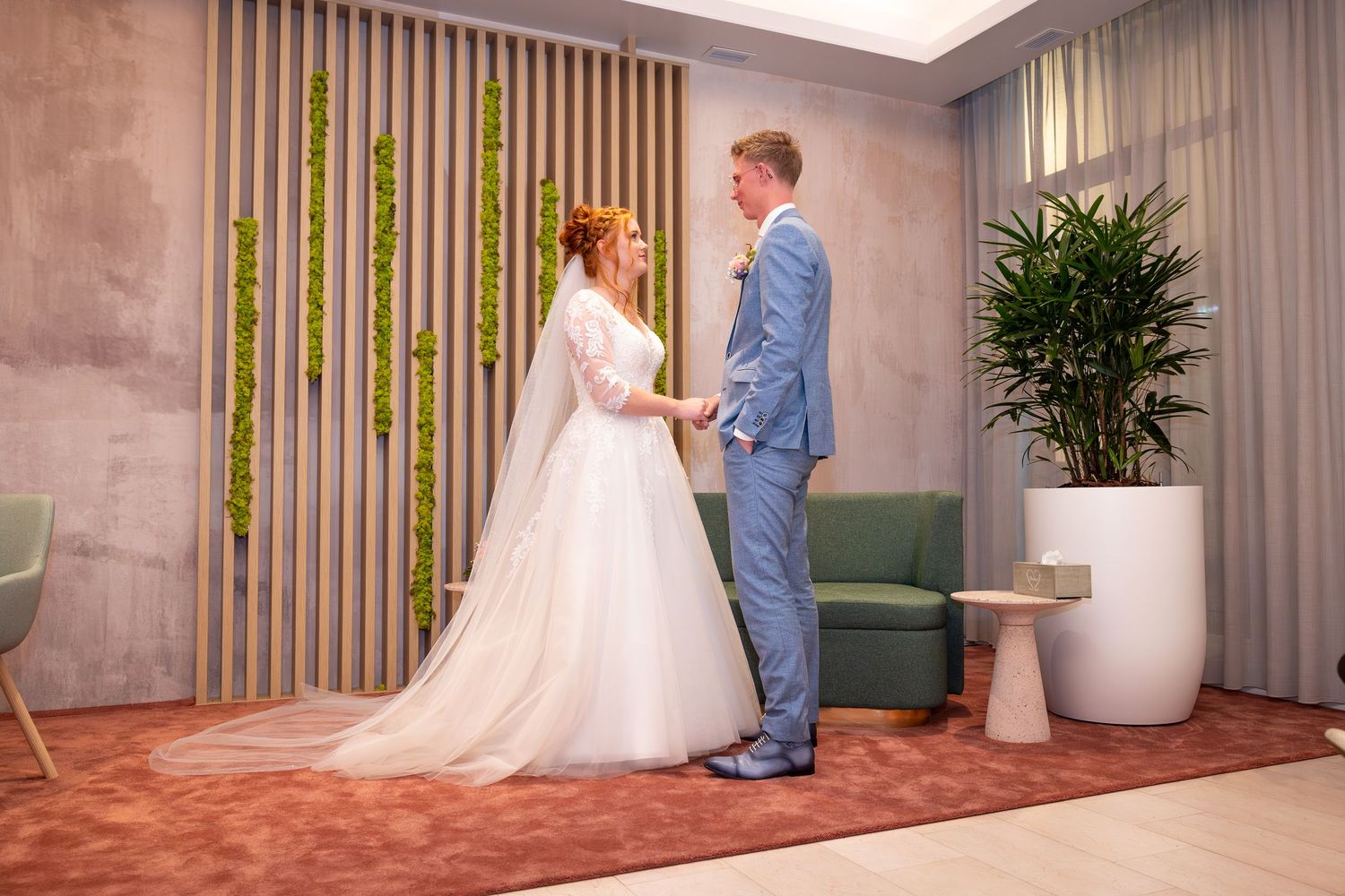A wedding couple holds hands in a modern room with bamboo accents and a potted plant.