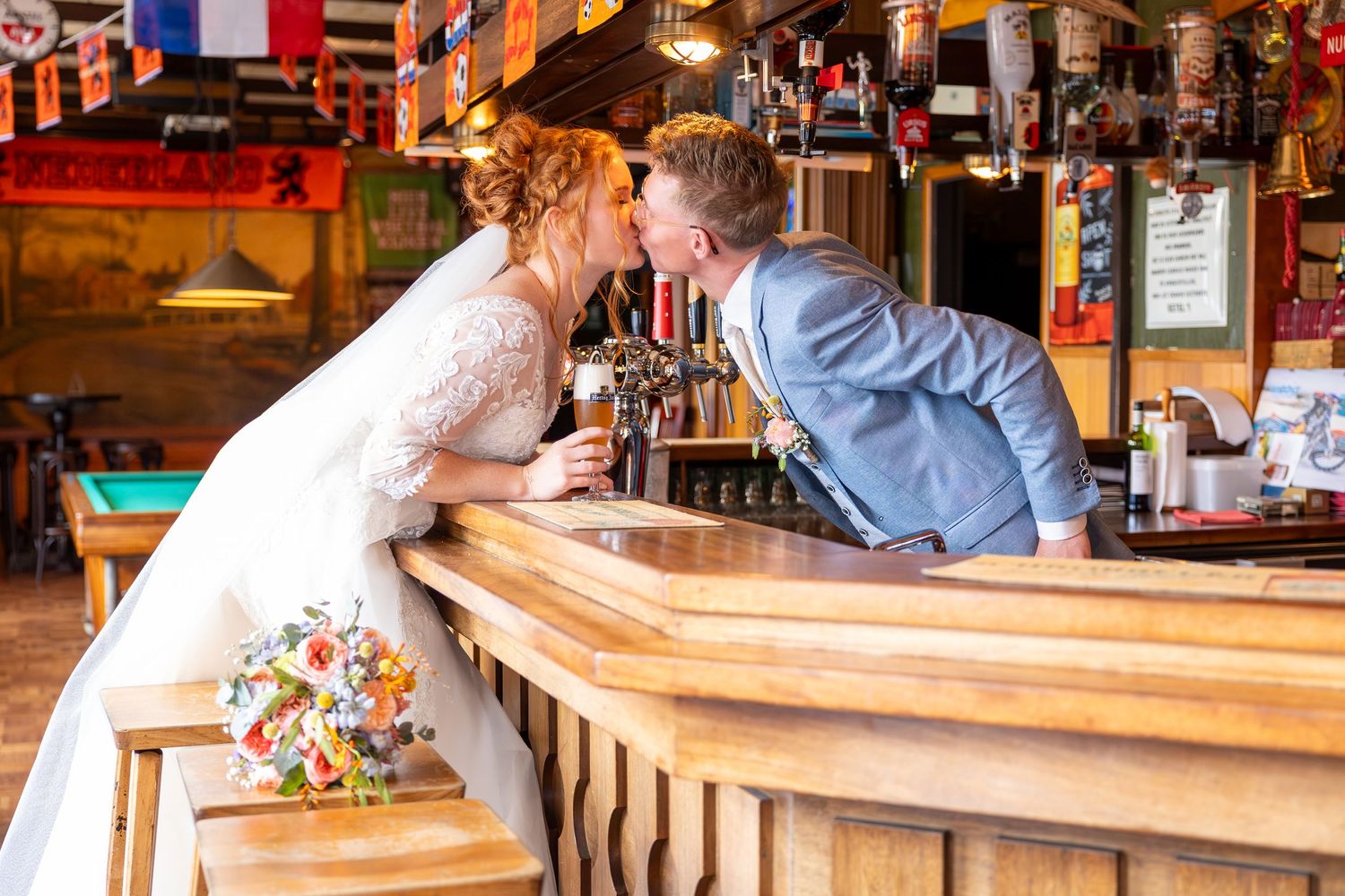 A couple in wedding attire leans across a wooden bar counter in a pub setting.