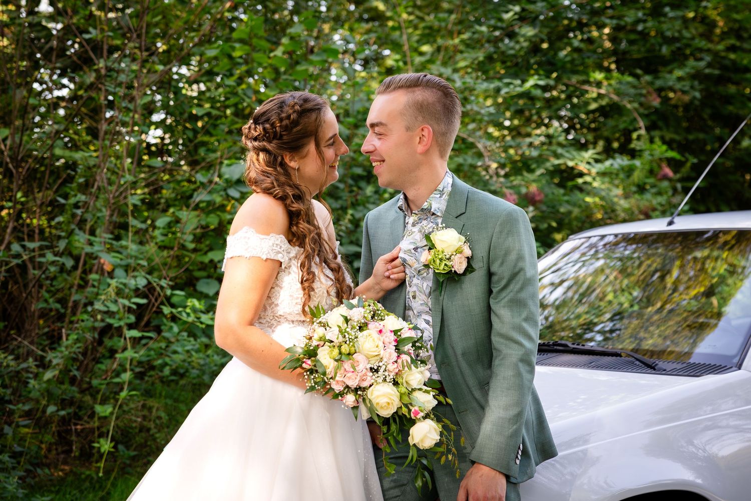 Een elegant bruidspaar in een grijs pak en een witte jurk poseert met een oldtimer in een bosrijke omgeving.