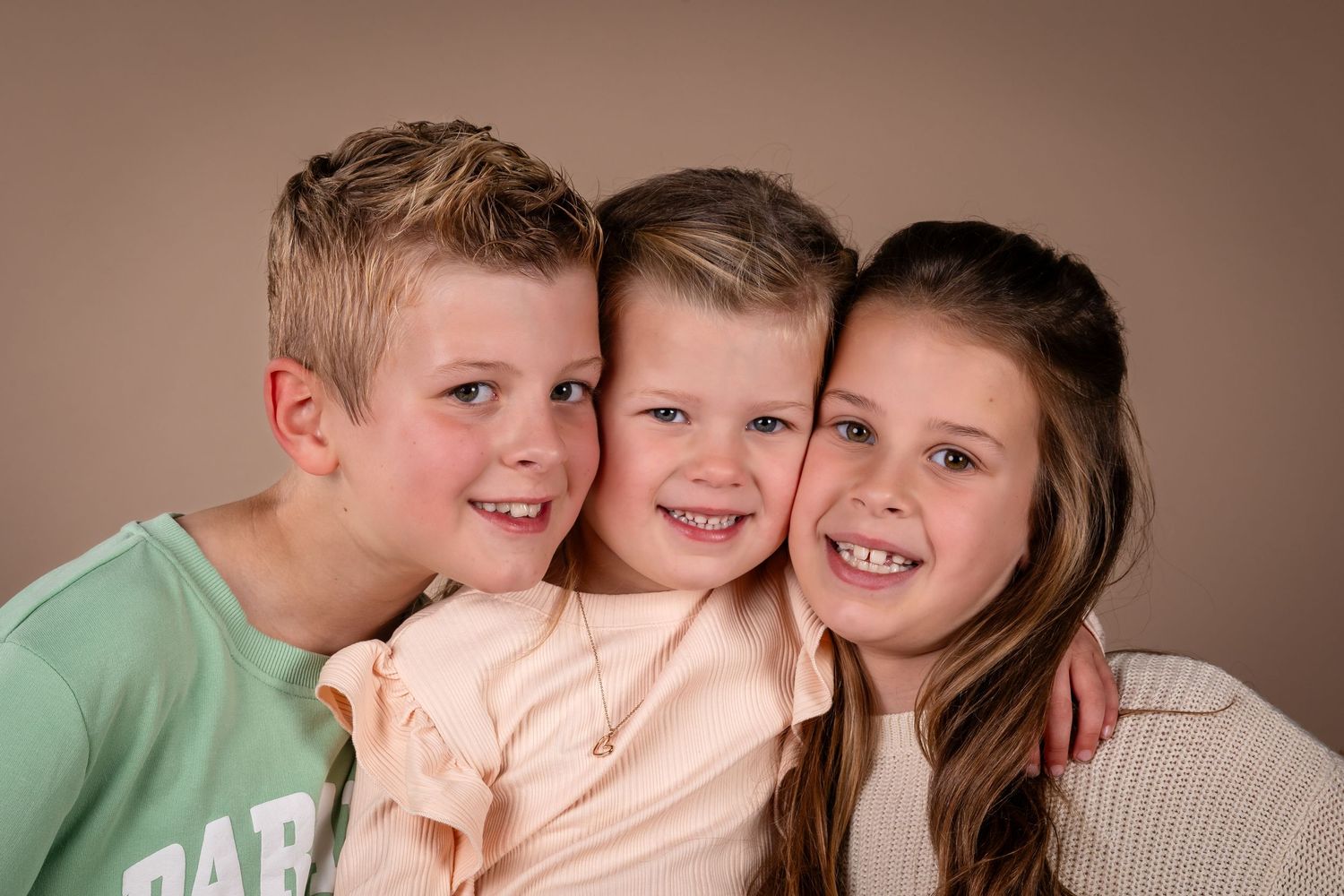 Three young siblings pose together against a brown backdrop wearing casual clothing and bright smiles.