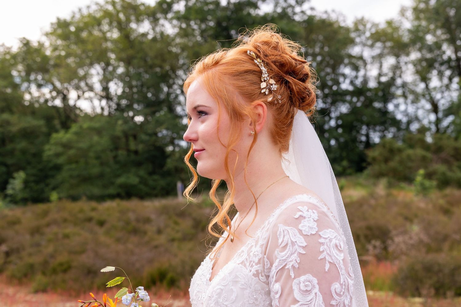A bride in a lace wedding dress with flowing veil stands in a natural heathland setting.