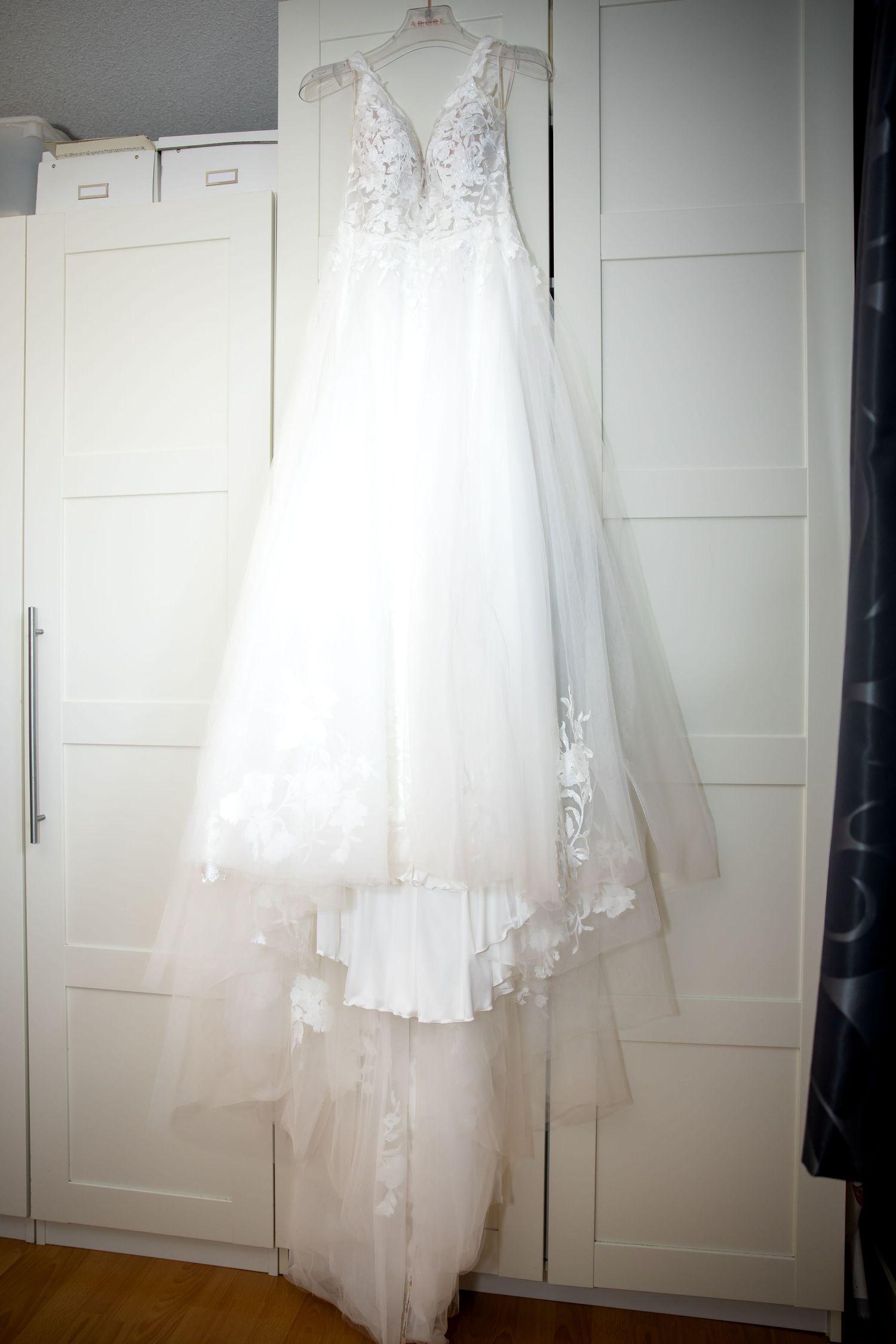 Een witte trouwjurk hangt elegant tegen witte kastdeuren, waardoor de delicate kanten details en de zwierige rok goed te zien zijn.