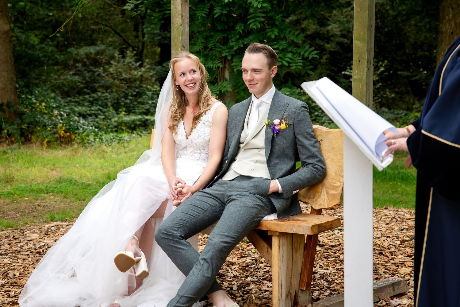 Een huwelijksceremonie vindt plaats in een rustiek houten paviljoen in de buitenlucht, omgeven door weelderig groen.