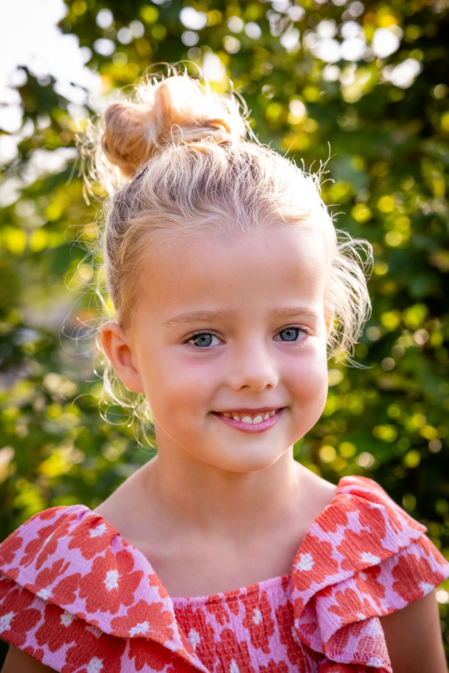A child in a red and white ruffled dress smiles in a sunny outdoor setting with bokeh-lit foliage in the background.
