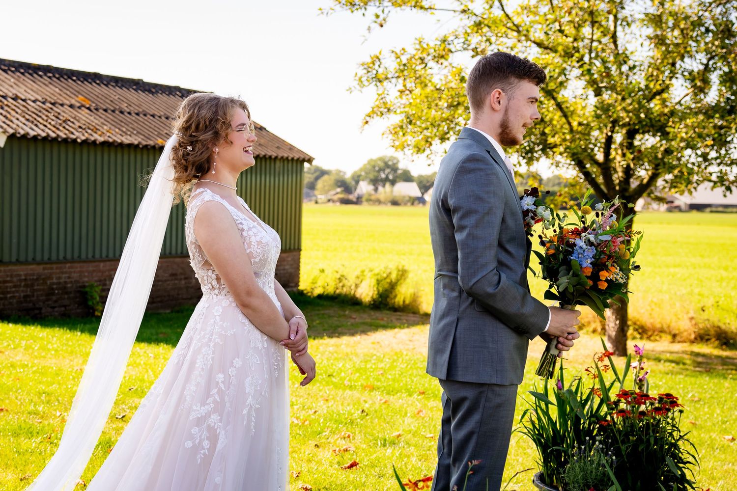 A wedding couple stands in a sunlit field next to a rustic barn with golden fields in the background.
