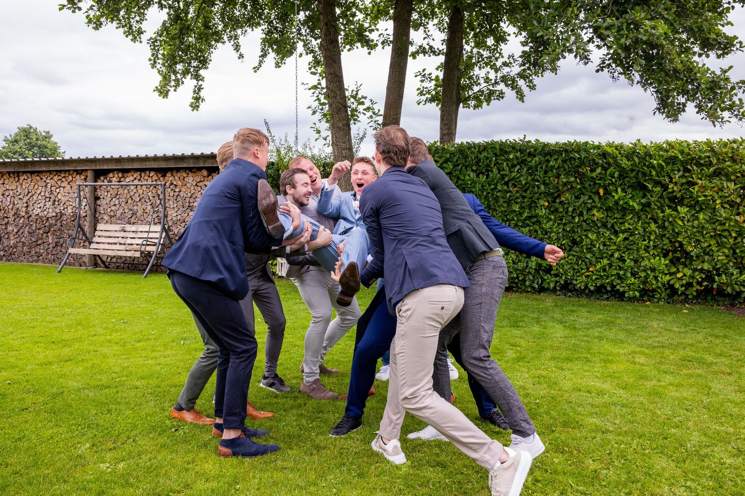 Groomsmen engage in playful group tussle on garden lawn during wedding festivities.