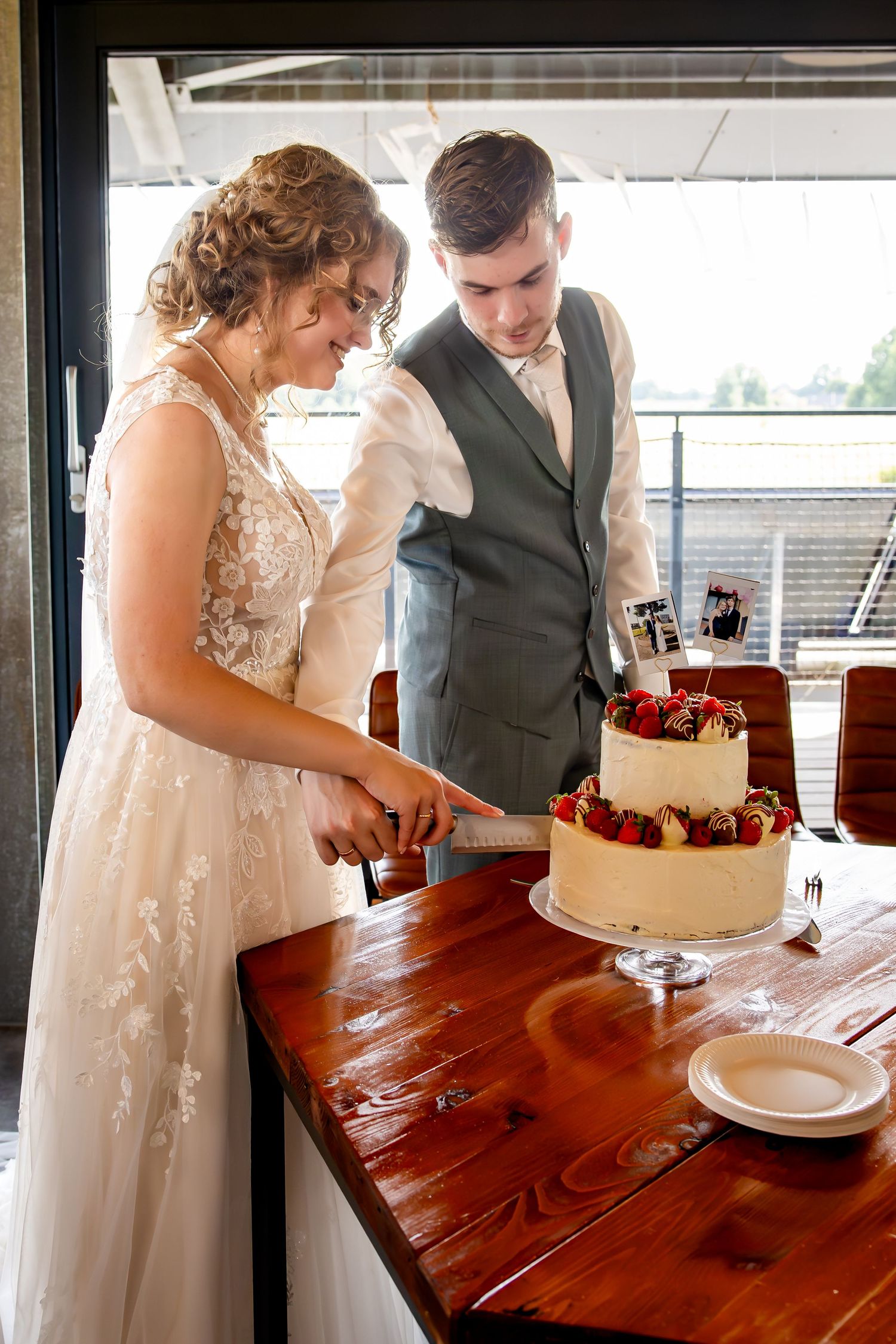 Wedding couple cuts their white wedding cake together at a rustic wooden table indoors.