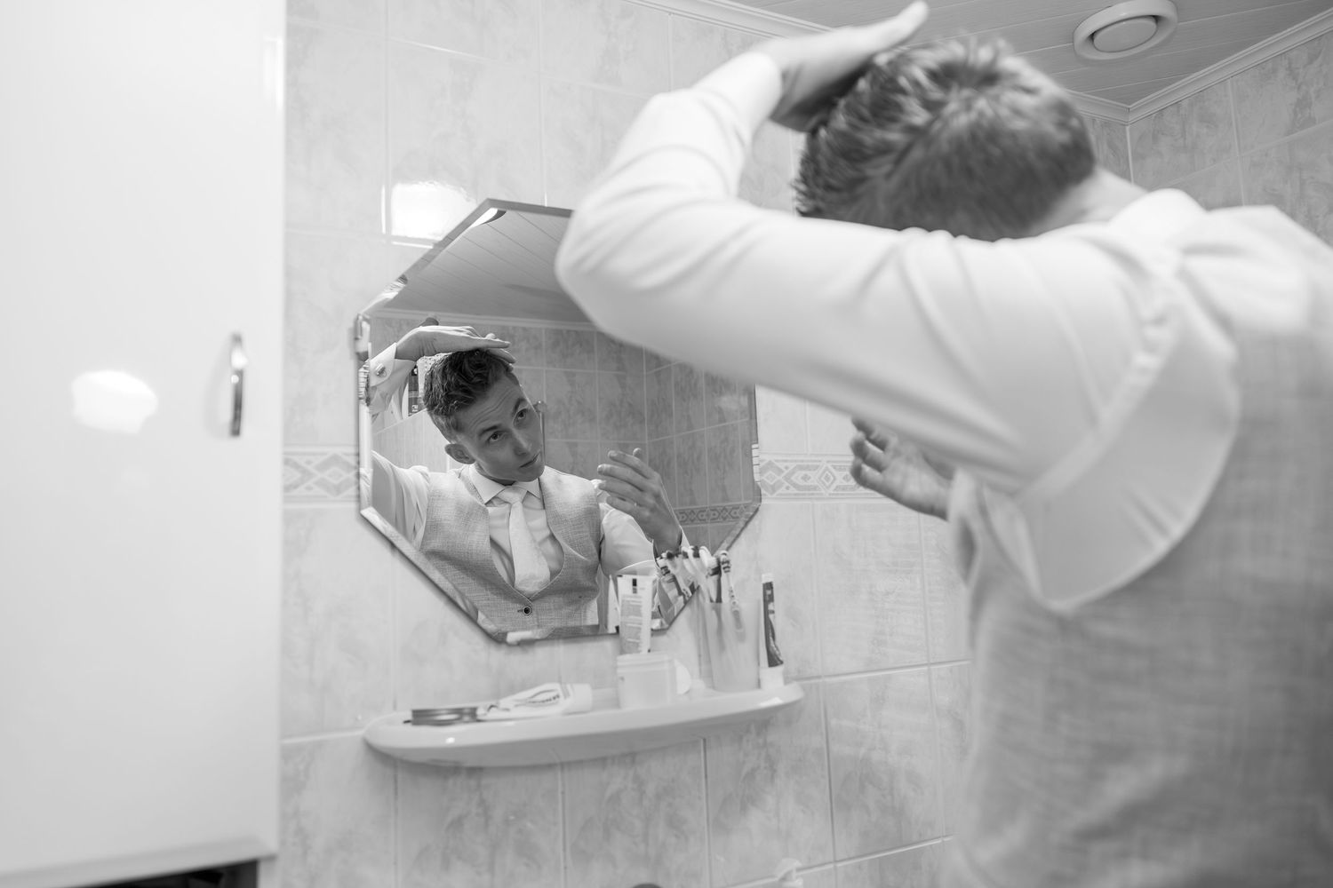 Black and white photo of person styling hair in bathroom mirror.