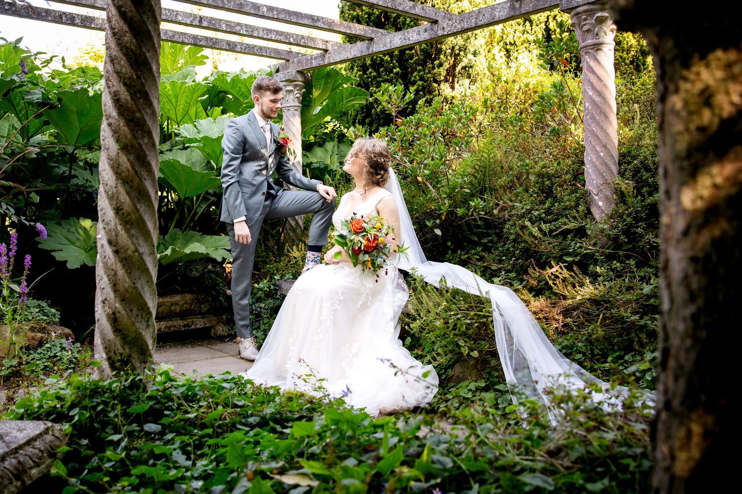 Wedding couple posing in a lush garden setting with natural sunlight filtering through trees.