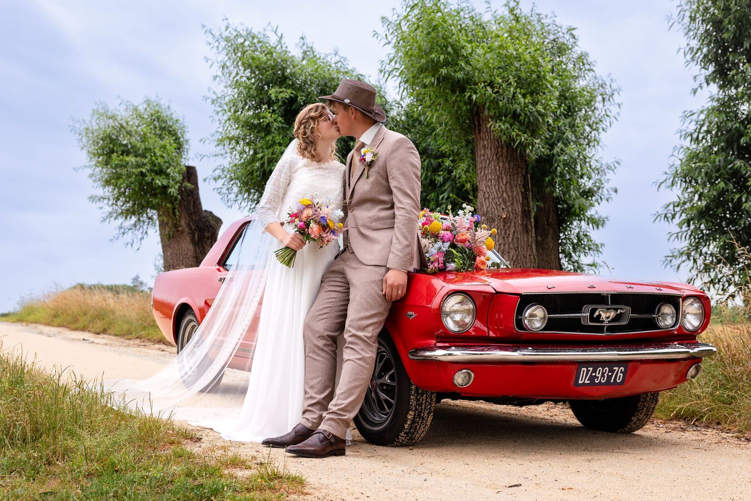 A wedding couple poses beside a classic red Ford Mustang on a country road.