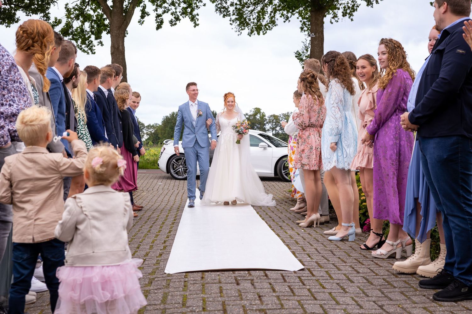 Wedding ceremony guests line both sides of a white aisle runner on a brick paved outdoor venue.