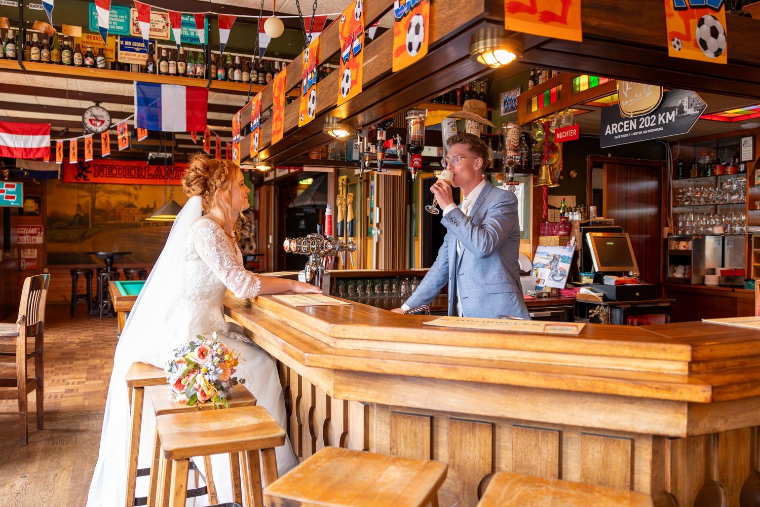 A couple in wedding attire interacts at a rustic wooden bar decorated with international flags.
