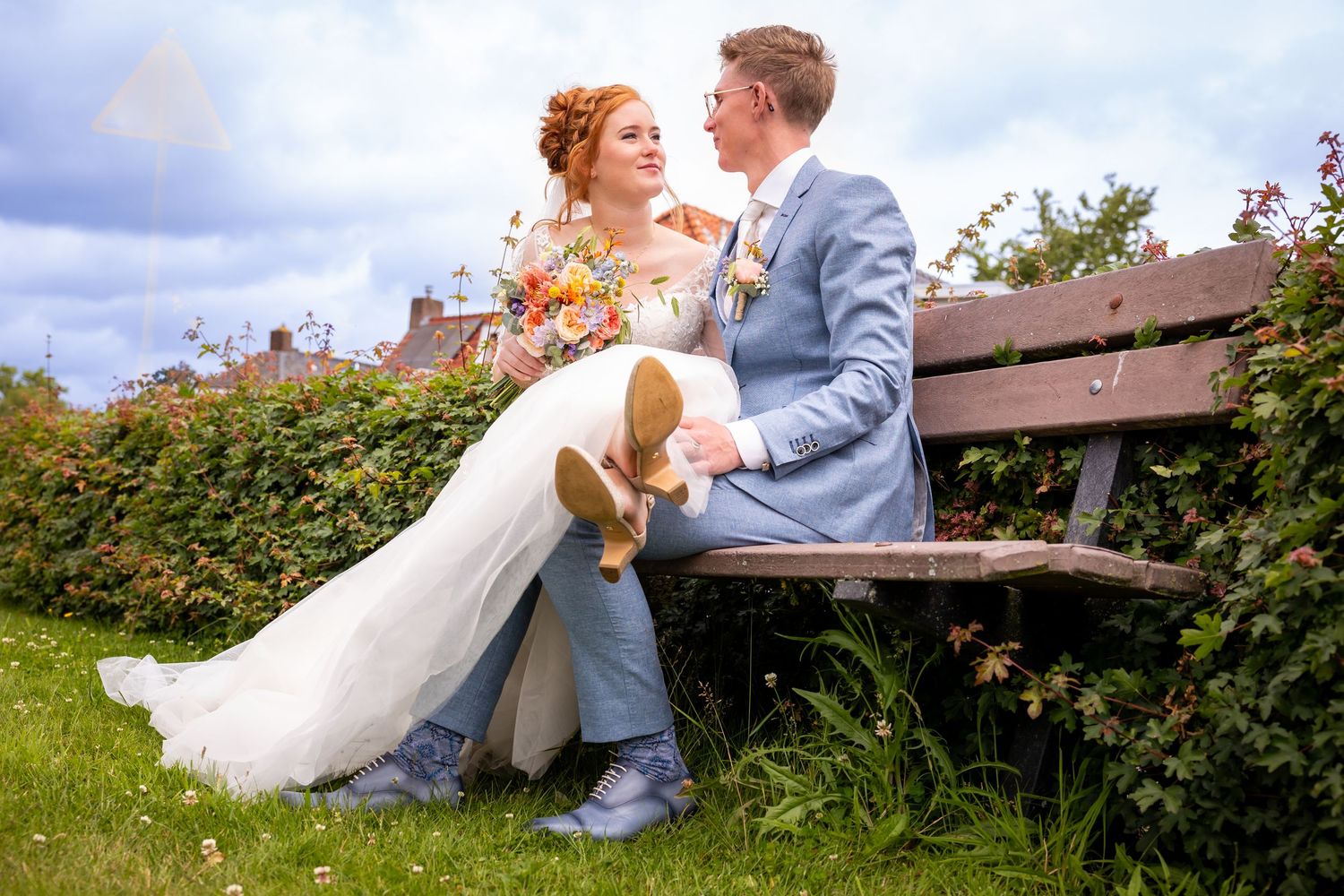 A couple in wedding attire sits on a wooden bench surrounded by greenery and flowers.
