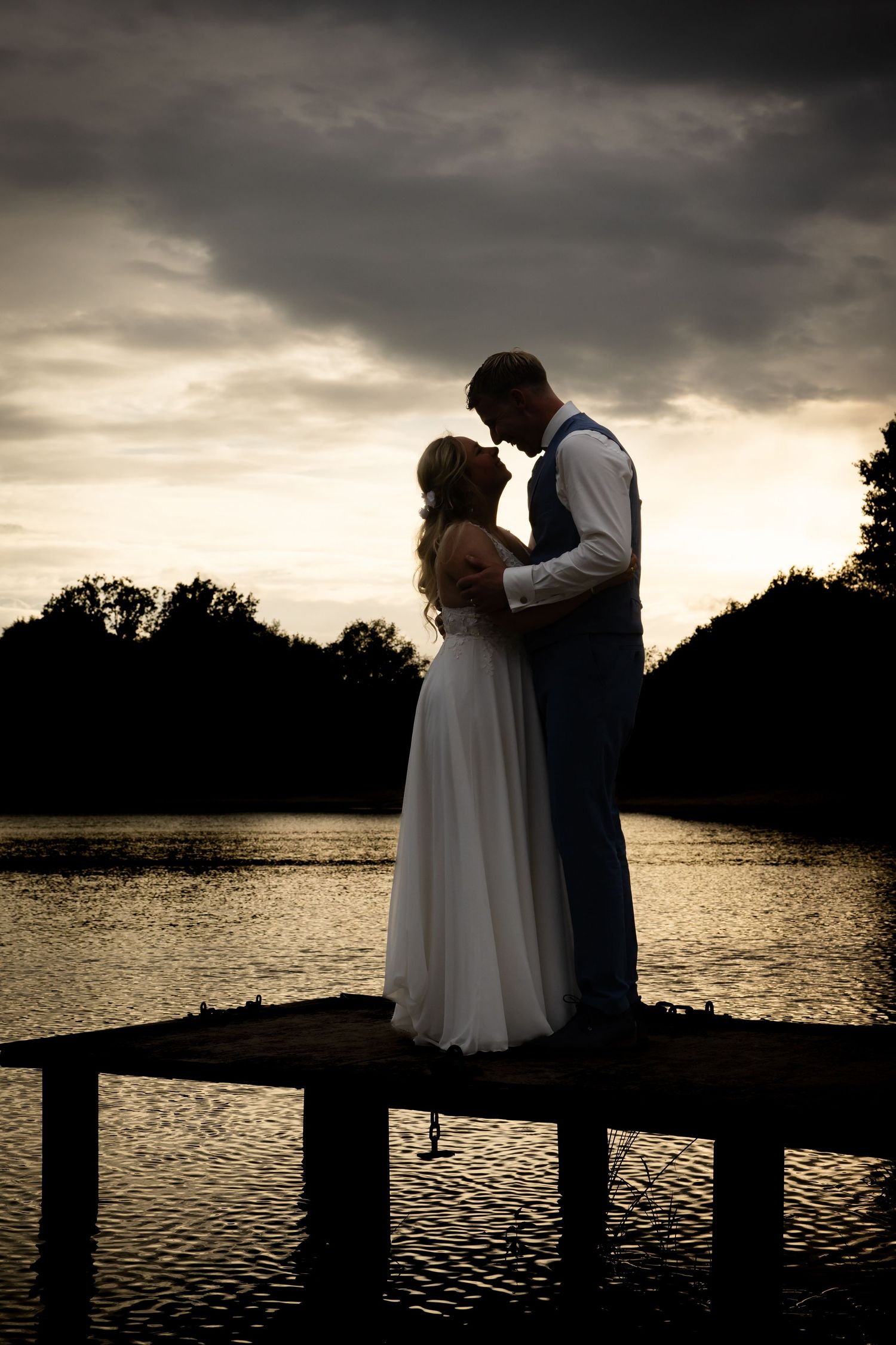 Silhouette of a bride and groom embracing on a dock at sunset with dramatic cloudy sky and lake backdrop.