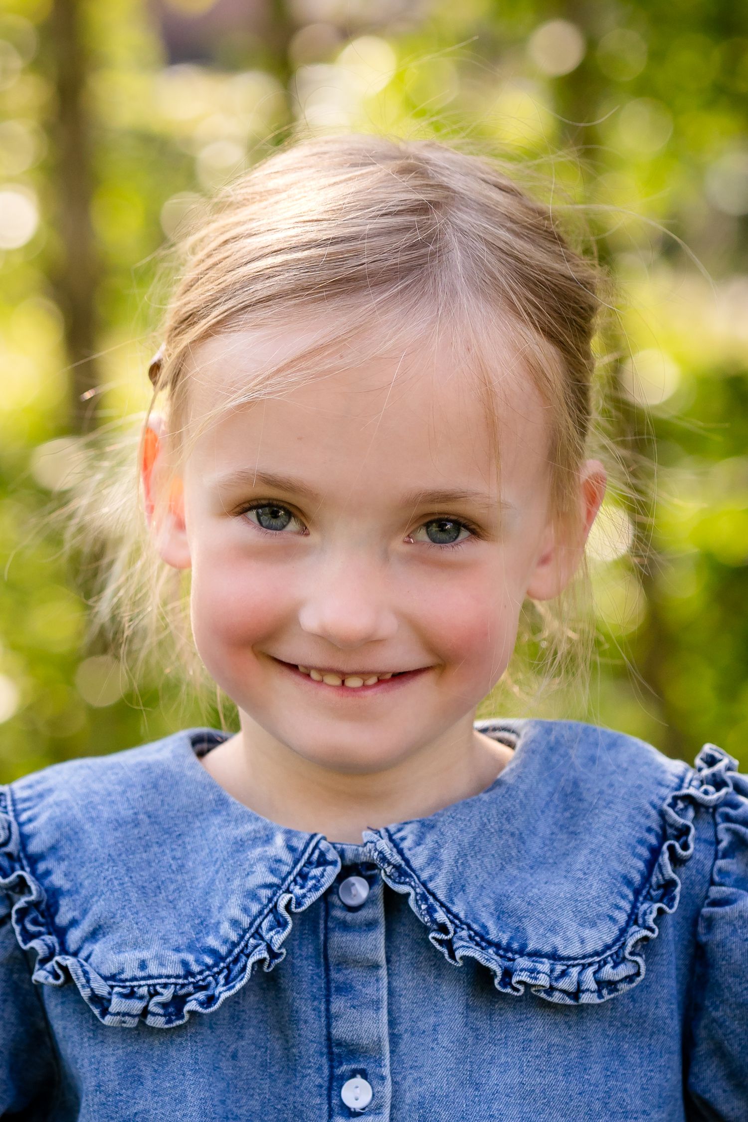 A young child wearing a blue denim dress with a ruffled Peter Pan collar smiles outdoors against a blurred green background.