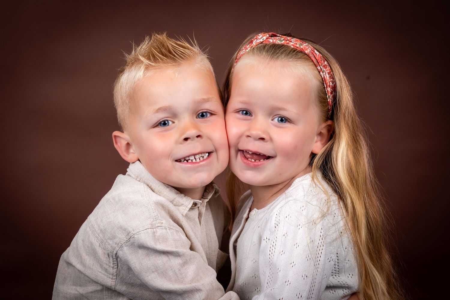 Two young children in white clothing pose together and smile warmly against a dark brown studio backdrop.