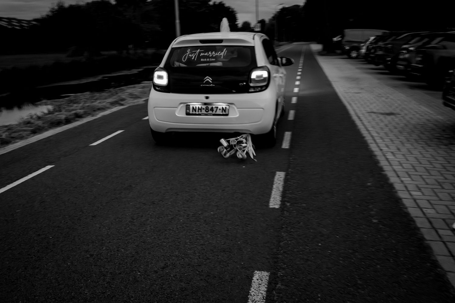Een zwart-wit foto van een de bruidsauto dat weg rijdt met het bruidspaar. er hangen blikjes achter de auto