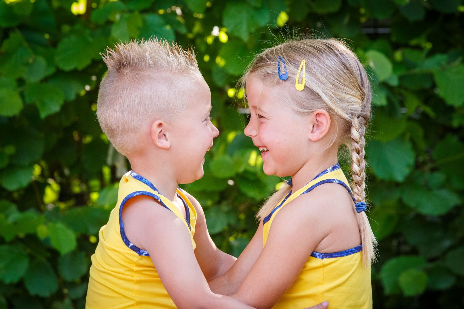 Two young children in yellow outfits share a sweet moment together while playing outdoors in front of green foliage.