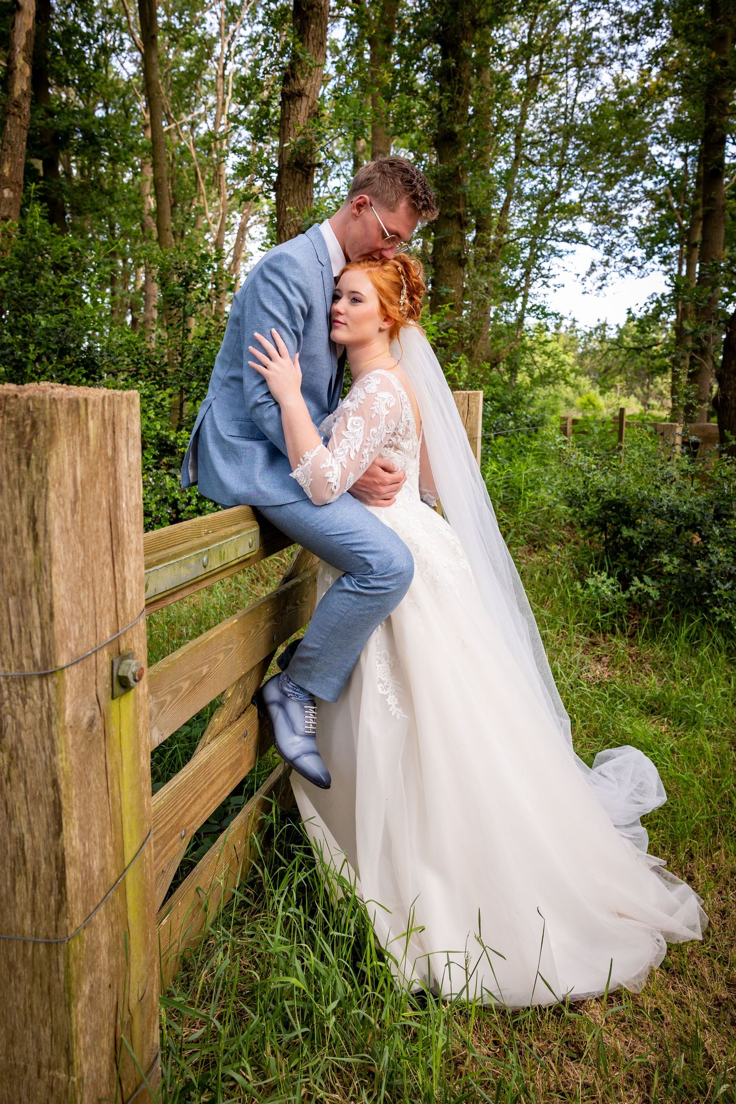 Bride in white gown and groom in blue suit share a romantic moment by a wooden fence in a lush green forest.