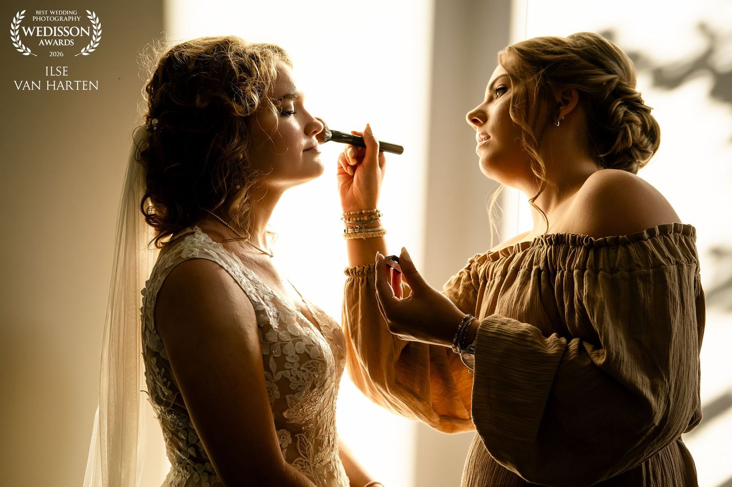 A makeup artist applies cosmetics to a bride in a golden backlit setting, captured by Ilse van Harten Photography.