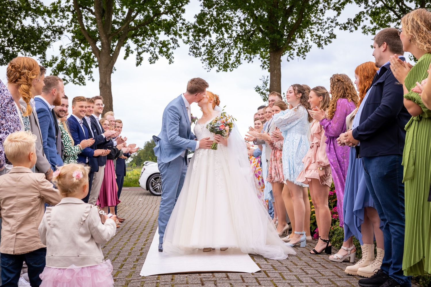 Wedding couple stands on white aisle runner surrounded by guests in colorful dresses and suits.
