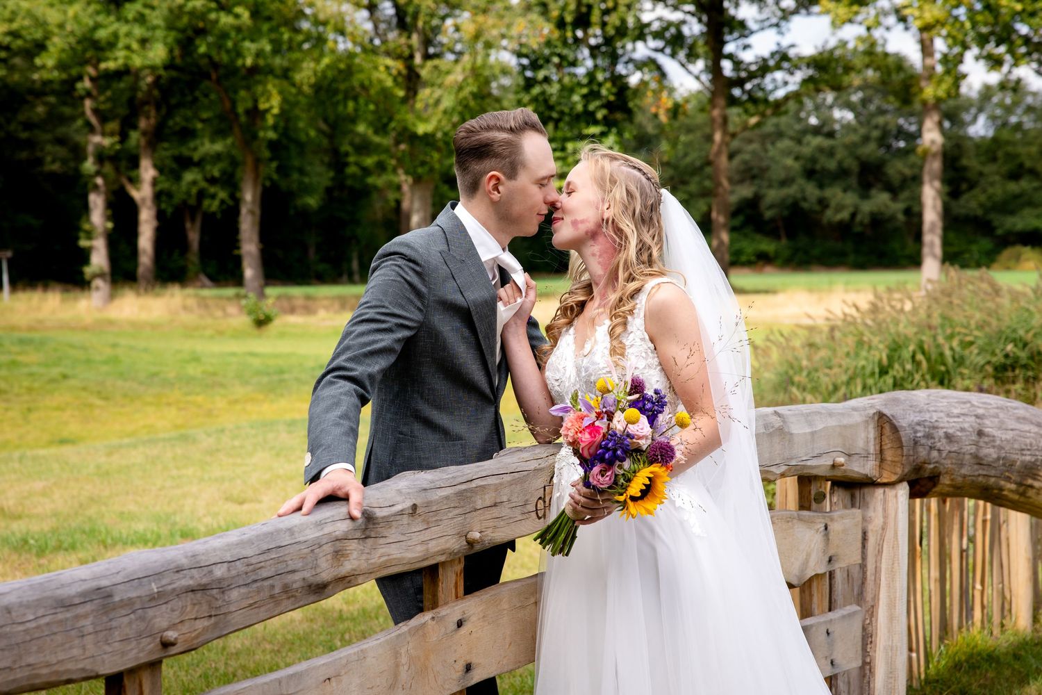 Romantisch trouwportret van een stel dat een teder moment deelt op een houten brug met een kleurrijk boeket.