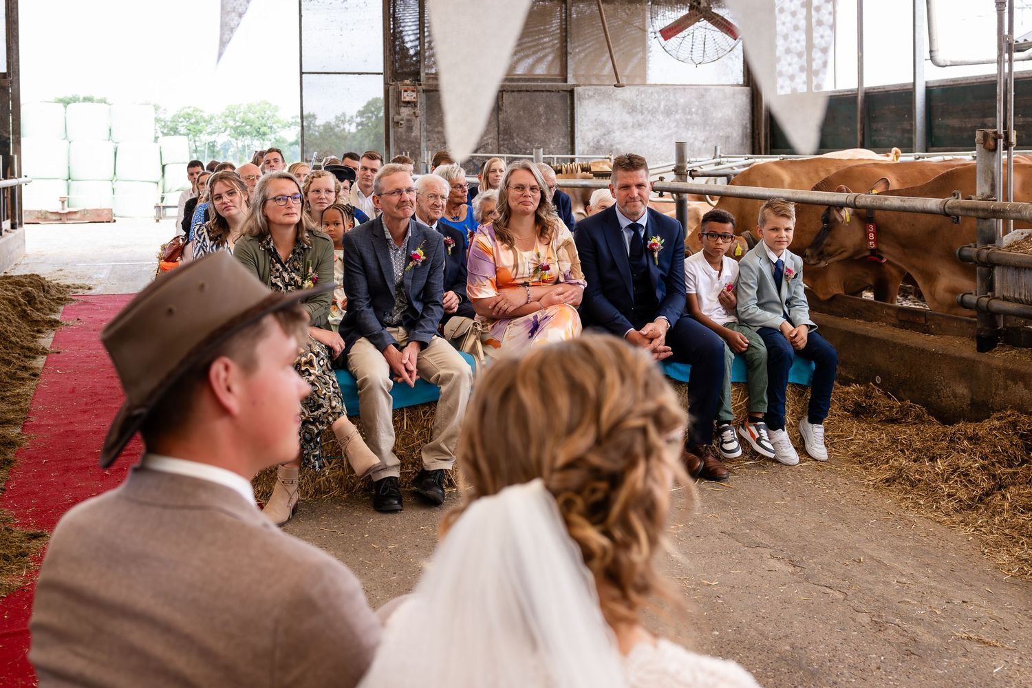 Rustic barn wedding ceremony with guests seated on hay bales along a red carpet aisle.
